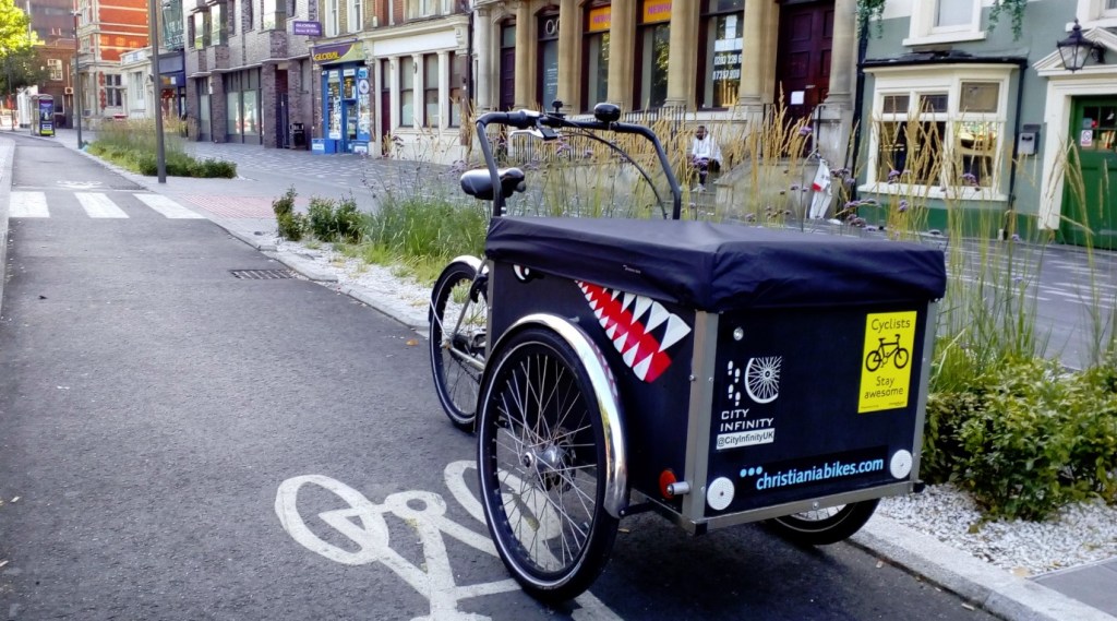A photograph of a black cargo-tricycle with a shark's face on the side, the City Infinity logo on the front and a yellow "cyclists stay awesome" sticker on the front. The trike is sat on a cycle track with lush planting to the right and a footway and shops beyond.