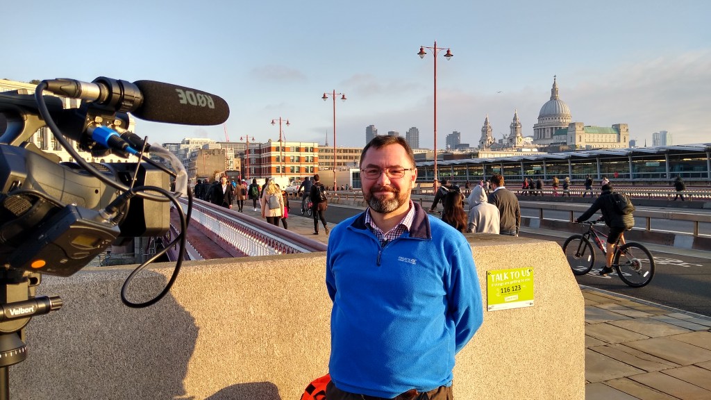 A photograph of Mark in a blue top. There is a video camera to the left, he is on Blackfriars Bridge with people walking and cycling behind him. St. Paul's Cathedral can be seen in the distance.