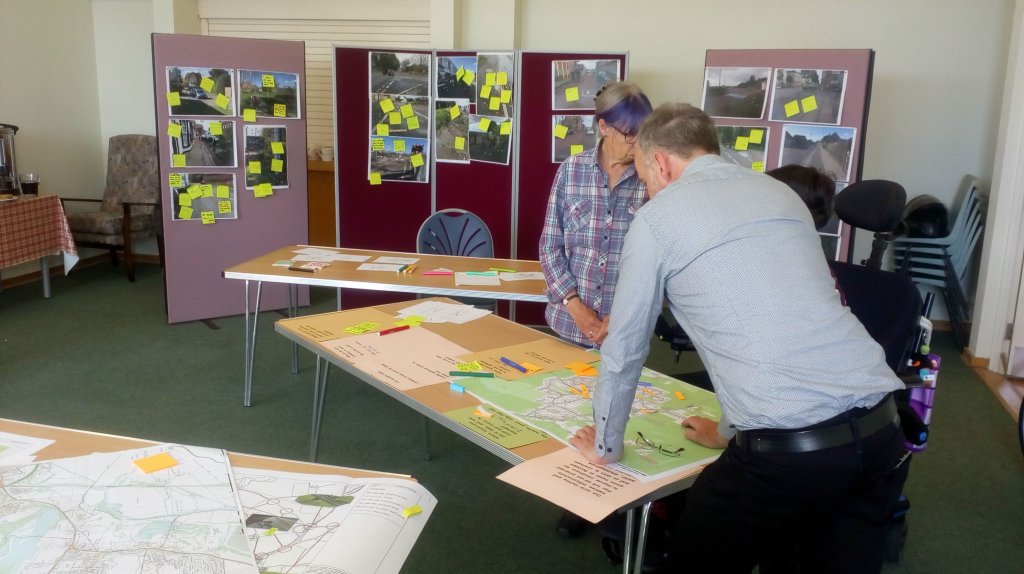 A room with three presentation boards covered in photos with Post-it notes on them. There are two tables with plans, pens and paper and a man and a woman are at one table looking a a plan.