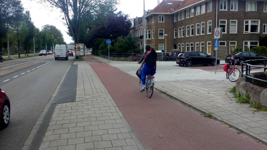 A view of a red cycle track with a road to the right and a footway to the left. A side road joins from the right and drivers have to negotiate ramped kerbs.
