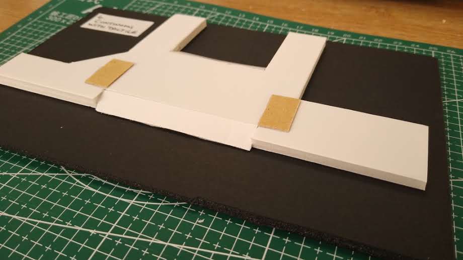 A model made from foam sandwiched between paper. The model is from a slightly elevated position and it depicts a continuous footway over a side street at a T junction. The road is black, the footway is white and there are rectangles of buff material each side of the crossing of the side road to show tactile paving.