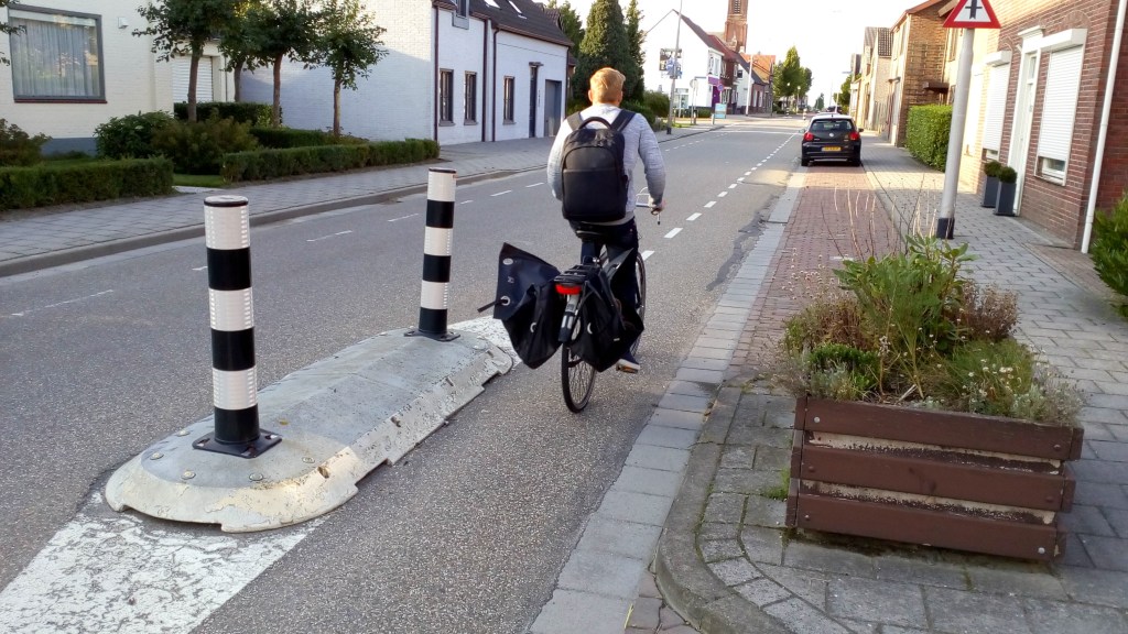A man cycles on the right hand side of a village road separated from traffic by a short traffic island.