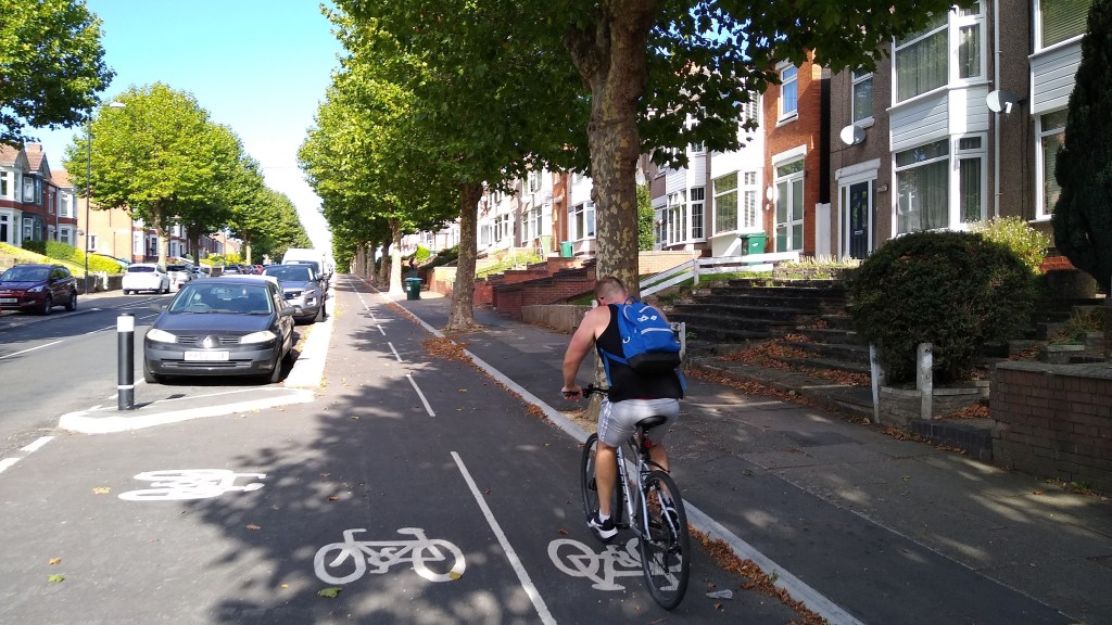 A cyclist rides on a cycle track along a residential street lined with trees and houses. He is going up quite a steep hill.