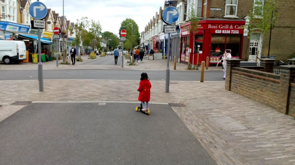 A child in a red coat rides a scooter down a street lined with shops, featuring directional and no entry signs. Several pedestrians are visible in the background.