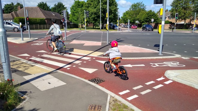 A parent and child riding bicycles on a red cycling path at a road intersection, with traffic lights and trees in the background.