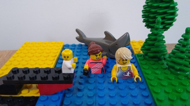 A colorful LEGO scene depicting two minifigures swimming in blue water, with a grey shark and a smiling baby on a half finished bridge. A shark fin is visible in the water, along with green trees in the background.