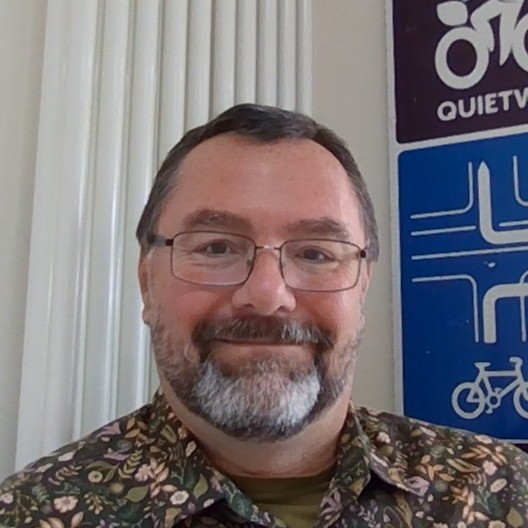 Mark, a middle aged white male with a greying beard, short hair and glasses wearing a brown flowery shirt.