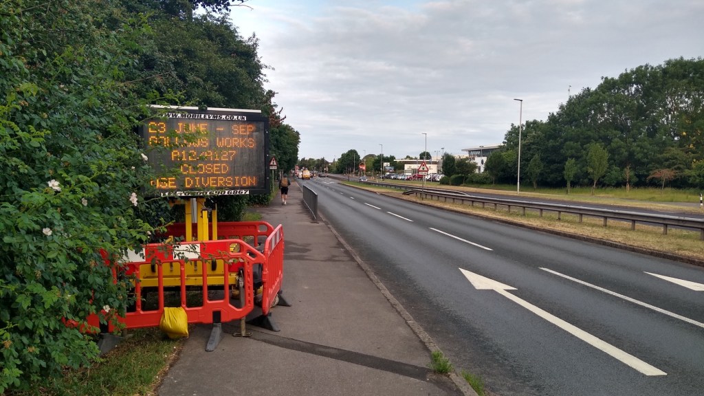 A mobile VMS sign on a yellow trailer with orange barriers round it, half in a verge with bushes and half on a shared path with a dual carriageway immediately to the right.
The sign says in orange lit letters "23 June - Sep Gallows Corner Works A12/A127 closed use diversion" and that is partially obscured by bushes.