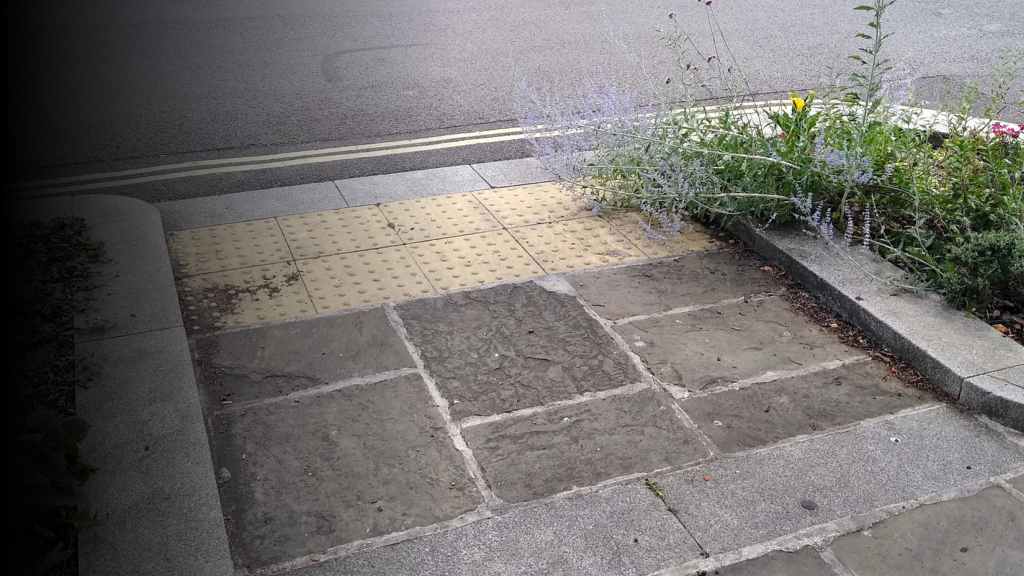 A dropped kerb viewed from the footway. It slopes gently towards the road with stone paving and two rows of buff tactile paving, about 2 metres wide. There are kerbed, planted areas on both sides.