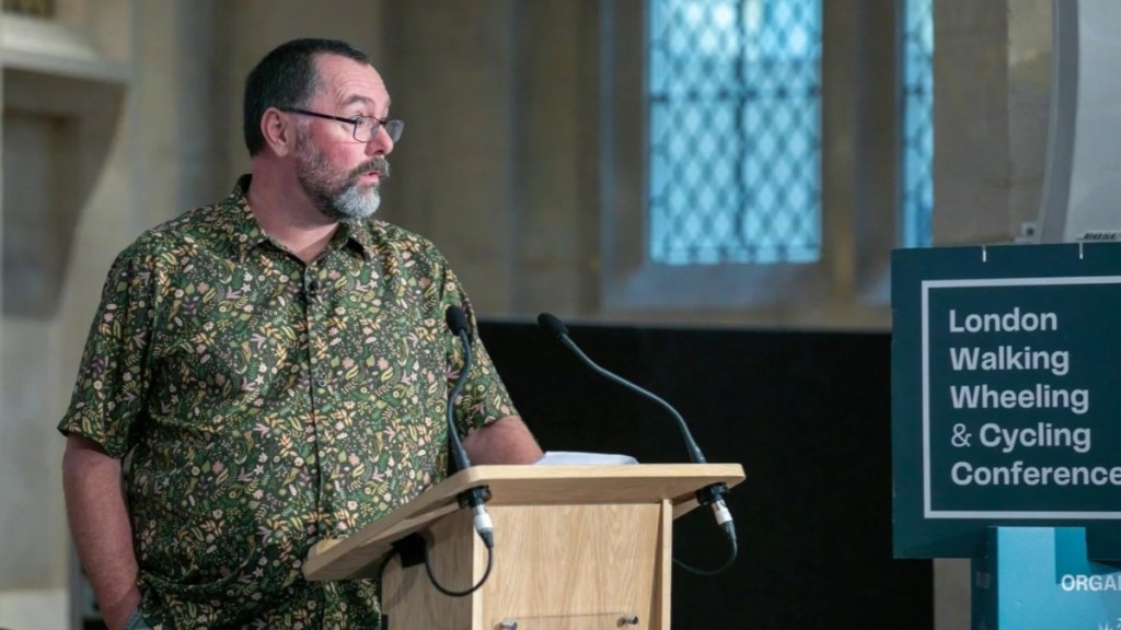 Mark, a middle aged white man with short dark hair, a grey beard and glasses is at a lectern talking with a serious look on his face.